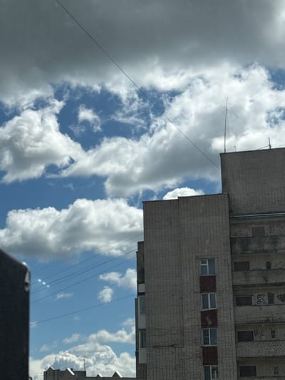 The image depicts a view of the sky and parts of a building, oriented such that the sky occupies the majority of the frame. The sky is filled with a mix of dark, heavy clouds on the left and bright, puffy white clouds against a blue sky on the right. Several thin, dark wires or cables crisscross the sky.
The building, visible in the upper right and extending down the right side of the frame, appears to be an apartment complex or similar multi-story structure. It is constructed of light-colored brick or tile and features rows of windows. Some windows are dark, while others appear to be glass panes, and a few are boarded up or covered with reddish material. Graffiti is visible on some sections of the building's facade.
The overall impression is that of an overcast but potentially clearing day, possibly late morning or early afternoon given the lighting on the clouds. The location is identified as Radkevshchina, Russia, suggesting an urban or suburban setting. No people are visible in the image. The scene is static, with no discernible activity taking place. The mood is neutral, focusing on the interplay of clouds, sky, and architecture.