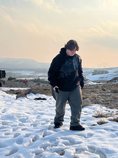 The image displays a snowy, mountainous landscape located in Talfita, Syria. In the foreground, a single individual stands slightly left of center, facing forward with their head angled downwards. This person possesses medium-length dark hair and is attired in a dark hooded sweatshirt featuring a lighter-colored pattern on the chest, dark grey sweatpants, and dark shoes or boots. A dark-colored object, likely a mobile phone, is partially visible in their right hand, held against the thigh. The ground surrounding the individual is covered in packed snow, exhibiting multiple footprints.
The immediate environment consists of uneven terrain with snow patches interspersed with exposed dry earth and sparse, dormant vegetation. In the midground to the left, a concrete or stone structure is visible, along with a prominent red cylindrical object, possibly a water tank. Further to the right midground, a dark-colored building is partially visible amidst the snow. The distant background reveals a series of rolling, snow-dusted hills or mountains beneath a pale, warm-toned sky with minimal cloud cover. Two metallic lattice-frame power transmission towers are discernible on the horizon line toward the right side of the frame.