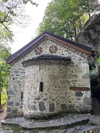 The image depicts a stone building with a gabled roof, likely a chapel or small church, situated in an outdoor, possibly wooded, area. The stonework of the building is varied, with different sized stones and some decorative elements, including circular designs and possibly crosses near the roofline. The roof has a dark trim. To the right, there is a stone-paved area or walkway. A sign with Cyrillic lettering is visible on the wall of the building. The environment includes trees and vegetation, and part of a large rock or cliff face is visible at the top of the frame. According to information found on the internet, this may be the Church of the Ascension of God in Govedartsi, Bulgaria.