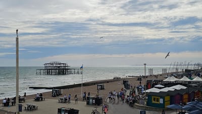 This is a high-angle, wide shot of Brighton Beach in Brighton, United Kingdom, on a cloudy day. The image captures the Brighton Pier's ruined structure in the distance, with the sea stretching out to the horizon. In the foreground, a paved promenade runs along the pebble beach, dotted with people enjoying the seaside. Beach huts in various vibrant colors line the promenade, and several boats are visible on the sand. The sky is filled with scattered clouds, and a seagull is captured in flight. The scene depicts a lively beach atmosphere with a mix of leisure activities and seaside infrastructure.