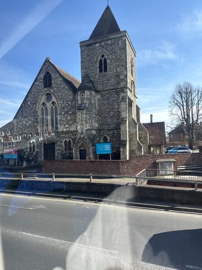 The image displays St. Thomas's Church in Salisbury, United Kingdom, viewed from across a road under a clear blue sky. The church, constructed primarily from grey stone and flint with lighter stone dressings, features a prominent square tower topped with a dark, pointed spire. Gothic-style arched windows, some with louvres, are visible on the tower and the main body of the church. The main section of the building has a steeply pitched, dark tiled roof. An additional building with a red-tiled roof adjoins the church to the right.
In the foreground, a paved road with a double white line runs horizontally, exhibiting reflections and glare from bright sunlight, suggesting the photograph was taken through a window. A low, dark grey concrete barrier topped with a light-colored metal railing separates the road from a narrow paved footpath. Beyond this, a reddish-brown brick wall encloses the church grounds.
Two light blue signs are situated on the church grounds behind the brick wall. The left sign displays "Welcome" and potentially service times. The right sign reads: "someone's FAITH restoring HOPE rooted LOVE."
In the background to the far right, a tall, bare deciduous tree stands against the sky. A second, smaller building with a red-tiled roof is visible behind the church, partially obscured by the tree, with several parked cars (including blue and white vehicles) visible nearby. No individuals are present in the scene.