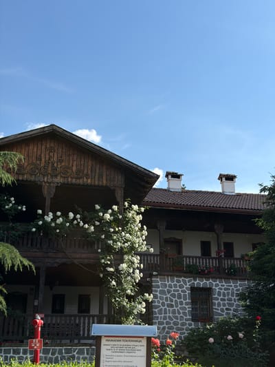 The image shows a building with a wooden upper story and a stone-clad lower story. There is a covered balcony on the upper level with a wooden railing, adorned with a large white climbing rose bush. The roof is tiled, and two white chimneys with dark caps rise from it. The building is set against a bright blue sky with scattered white clouds. In the foreground, there is a red fire hydrant with a sign, and a white information board with text in Bulgarian. Red and pink roses are blooming in front of the stone wall of the building. The overall scene depicts a historic building with lush vegetation in an outdoor setting. The text on the information board translates to "Dear Pilgrims. The monastery is a holy place with... God's... blessed... Respect the holy place... and its surroundings." It also lists rules such as "1. Bow to the monastery with reverence and humility!" and "2. Dress modestly!"