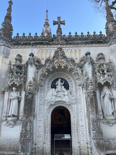 An ornate, white stone building facade with intricate Gothic detailing. Two statue niches flank a central arched doorway. Over the doorway, a round window is surrounded by sculpted figures of angels and a central figure. A large stone cross tops the structure. The sky is a clear blue. The facade is weathered and textured with moss and age. A spire with metal details is visible next to the cross.