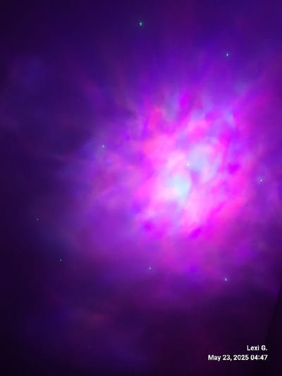 The image is a photograph of a purple and pink nebula-like projection displayed on a ceiling. The projection is centered in the image and appears to be made with a projector. There are no visible people or objects in the image, and the location appears to be an indoor space. The image is taken in Lexington, Kentucky, United States, based on the provided coordinates. The time and date are recorded in the bottom right corner of the image as "May 23, 2025 04:47." The image was taken by someone named "Lexi G." The environment is dimly lit with only the projection providing any illumination.