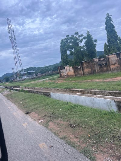 In the city of Kona Garu, Nigeria, a telecommunications tower stands tall on the left side of the frame, its red and white lattice structure reaching towards a cloudy sky. In the foreground, a paved road with yellow lane markings runs diagonally from the bottom left corner. Adjacent to the road is a grassy verge with a concrete drainage channel running alongside it. Beyond the drainage channel, a weathered, ochre-colored wall with occasional graffiti runs horizontally across the midground. Several tall, dark green trees stand behind the wall. In the background, a cluster of traditional buildings with thatched roofs are visible, nestled amongst lush green vegetation and rolling hills. The overall atmosphere is overcast and slightly humid. There are no people visible in the image.