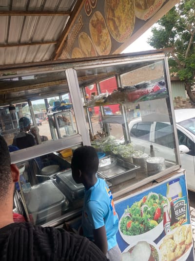 The image shows a food stand with a glass front in Adenta Municipality, Ghana. The stand is made of metal and has a tin roof. It is located in a bustling street with traffic. On the roof of the stand is a sign advertising "Jain Rice" and fried chicken. The stand has a variety of food items for sale, including salad, fried chicken, and rice. A young boy in a blue shirt is standing in front of the stand looking at the food. Another adult, partially obscured, is standing to the left, looking at the boy and possibly at the food. Both are standing at the stand, waiting to be served. The stand is lit up with bright lights, making it an attractive and inviting place to buy food. The street outside the stand is busy with traffic and pedestrians.