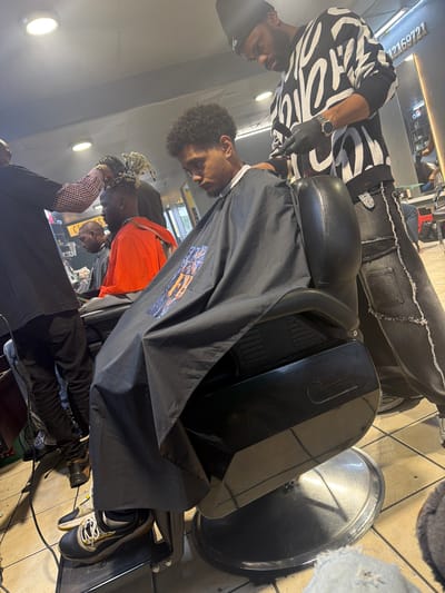 The image portrays an active scene inside a barbershop in Blouberg, South Africa, capturing a haircut in progress.
In the foreground, a barber with dark skin and a beard is visible on the left. He wears a black beanie featuring "AIR" text, a distinct black and white patterned long-sleeved top, black gloves, and a wristwatch. He is focused on cutting the hair of a client seated in a large, black leather barber's chair, holding clippers or a trimmer in his gloved hand. His attire includes dark denim pants with frayed seams.
The client, a young male with dark, curly hair, is draped in a black barber's cape that displays a colorful orange graphic design. His feet, clad in yellow, black, and white athletic shoes, rest on the chair's footrest.
In the background, other individuals are present within the barbershop. One person with dreadlocks or braids, wearing an orange long-sleeved shirt over a patterned inner shirt, is partially visible. A large mirror reflects parts of the interior, including another person and a sign displaying the numbers "2169721", with partial text above that possibly reads "HAIR". The floor is tiled with light-colored squares, and various pieces of equipment, including a large silver basin and electrical wires, are scattered around. Circular light fixtures illuminate the indoor space. The scene depicts a lively and functional barbershop environment during business hours.