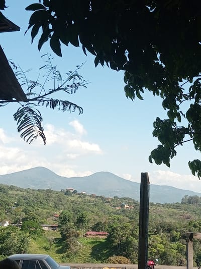 The image shows a landscape view in Honda, Venezuela. In the foreground, foliage partially obscures the sky. A road is visible, with a car and a motorcycle parked nearby. The midground features a lush, green hillside dotted with houses. In the background, two mountains are silhouetted against a light blue sky with scattered clouds.