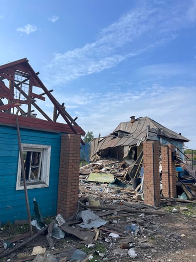 The image shows the aftermath of a destruction in Bilopillia, Ukraine (latitude: 51.139, longitude: 34.286). The scene is a residential area with two destroyed houses. The house on the left side of the image is a single-story building with a partially collapsed roof. The house on the right is a single-story building with a completely destroyed roof. The walls of both houses are made of wood and brick. The ground is covered in debris from the destroyed houses. There is a blue sky with white clouds in the background. The scene appears to be the result of a bombing or shelling. The debris is scattered in a haphazard manner, and there are no signs of any human presence. The image conveys a sense of destruction and loss.