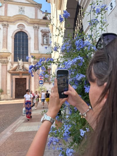 The image shows a person taking a photograph of a building with a smartphone. The building appears to be a church or a historic structure, with ornate architecture, a large arched window, and religious iconography. The person is holding the phone in landscape orientation, and the screen displays a preview of the photo being taken, showing the building. The person is wearing sunglasses on their head and a bracelet on their wrist. The foreground is filled with blooming blue flowers, which are also in focus in the lower part of the smartphone screen. In the background, other people are walking on a paved street. The scene is outdoors, suggesting a daytime setting. The weather appears to be clear and sunny. The overall mood is one of leisurely sightseeing and capturing memories. The image appears to be taken in Trento, Italy, given the visual cues of the architecture.