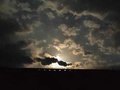The image depicts a dark silhouette of a rooftop against a cloudy night sky. The moon is partially visible through a break in the clouds, casting a soft glow that illuminates some of the cloud formations. The rooftop appears to be dark and flat, with a series of small, rounded objects visible along its edge, possibly skylights or decorative elements. The overall atmosphere is dramatic and serene, with the interplay of light and shadow creating a sense of mystery. The location is identified as Қарғалы, Kazakhstan, but no specific landmarks or context are provided in the image itself to further specify the exact location within the city.