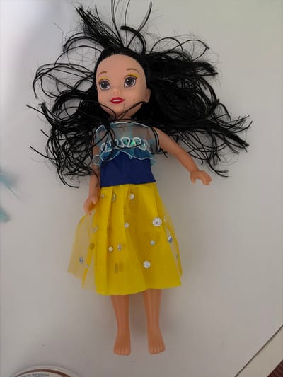 A doll with long black hair, a yellow and blue dress, and sparkly embellishments lies on a white surface. The doll is positioned horizontally, with its head to the left and feet to the right. The background is a plain, light-colored surface. There is a partial view of a round object with text on it at the bottom left of the image. The location is Limnia, Northern Cyprus.