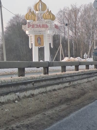 The image displays an outdoor scene depicting the entrance to the city of Ryazan, Russia, observed from a roadside vantage point near Turlatovo. A prominent white welcome sign for Ryazan stands centrally, featuring three golden, onion-shaped domes atop its structure. Below the domes, the city name "РЯЗАНЬ" is displayed in bold red Cyrillic letters. Beneath the text, a shield-shaped emblem depicts a standing figure, likely a warrior or prince, holding a sword.
In the foreground, a grey, multi-segment metal guardrail extends horizontally across the lower portion of the frame, separating the viewer from the area immediately surrounding the sign. The ground beside the road and behind the guardrail is covered with dirty, patchy snow and exposed brown earth. A paved asphalt road surface is visible in the lower right, partially obscured. The background consists of a dense line of bare deciduous trees under a light, possibly overcast, sky. Two utility poles are visible: one to the left of the sign and another to its right. A portion of the dark-colored cabin of a truck is visible on the far right edge of the image. No individuals are present within the scene.