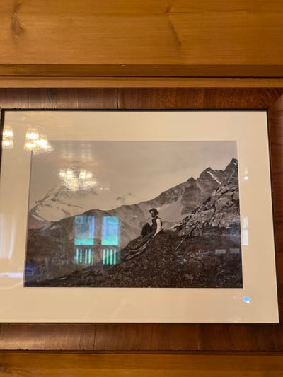 A framed black and white photograph hangs on a wooden paneled wall. The photograph depicts a rocky mountain landscape with a single person sitting on a rocky outcrop. The person, wearing a hat and vest, appears to be holding a pipe. In the background, rugged mountain peaks are visible. There are reflections on the glass of the frame, including what appears to be light fixtures. The lower part of the photograph shows a grassy slope. The image is located indoors, possibly in a home or establishment in Ленинское, Russia, given the presence of the wooden paneling.