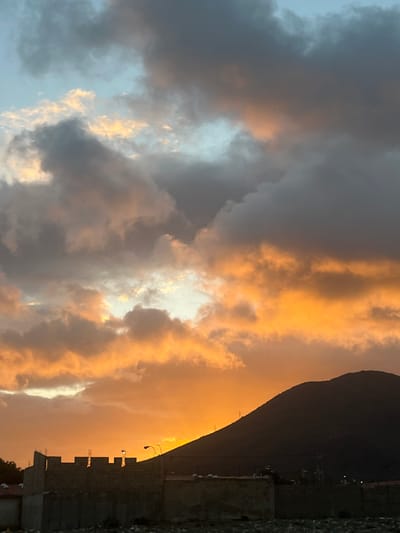 This image depicts a sunset over a mountainous landscape, likely in San Antonio, Venezuela. The sky is filled with dramatic clouds painted in hues of orange, yellow, and gray, with patches of blue sky visible. The sun appears to be setting behind a large, dark silhouette of a mountain. In the foreground, a partially constructed brick wall with visible rebar and some street lights suggest an urban or semi-urban environment. The overall impression is one of a rugged natural landscape meeting human development.