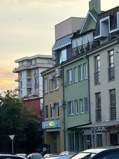 The media file depicts an outdoor scene featuring several multi-story buildings with varying architectural styles and colors. The buildings appear to be residential or commercial, with numerous windows and air conditioning units visible. Some of the buildings have balconies or terraces. In the foreground, a dark-colored car is partially visible, suggesting a street or parking area. Adjacent to the buildings, there are trees and foliage. A triangular traffic sign with a red border and a white interior is also present. The sky is visible in the upper part of the image, appearing overcast or at dusk, with faint cloud formations. A few birds are flying in the sky. The scene is oriented vertically, with the buildings extending upwards and the sky towards the top. The overall impression is an urban or semi-urban environment. There is text on a sign visible on one of the buildings; it is a circular logo with green and yellow elements and some text that is difficult to decipher. The image is a still photograph. The time of day appears to be late afternoon or early evening, given the lighting conditions and sky color. The weather appears to be mild. The location context provided is Khamkhi, Russia, which suggests a Russian urban or suburban setting.