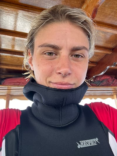 A woman with blonde hair and a nose ring poses in a black wetsuit with a high neck and red sleeves. The brand "NEOPRO" is visible on the suit. She has fair skin and light eyes. The background features wooden planks, possibly from a boat or structure. The image has been rotated, so the woman appears sideways.
