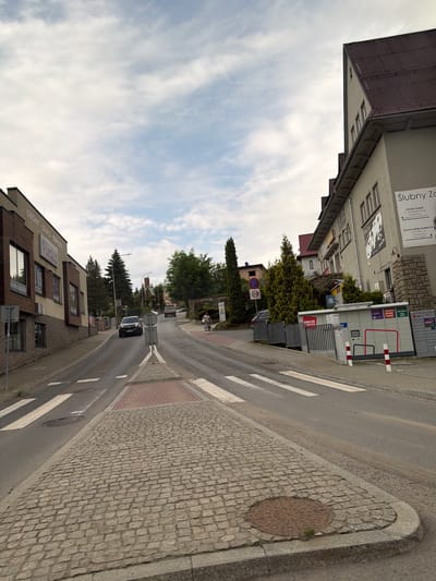 The image depicts a street scene in Limanowa, Poland. The foreground features a cobblestone sidewalk with a circular manhole cover. A pedestrian crossing with white stripes is marked on the asphalt road. In the middle ground, a black SUV is driving uphill. To the left of the road, a building with brown and beige facade displays signage for "papier biuro szkoła" and "Plastus." Further up the street, several trees are visible, including a tall evergreen tree and a cluster of cypress trees. On the right side of the road, a building with a dark red roof and light-colored walls has signage for "Ślubny Z...". Adjacent to this building is a row of parcel lockers and a fence. The sky is partly cloudy with a mix of blue and white. In the far distance, a truck is also driving uphill, and more buildings and trees are visible. A woman in a patterned dress and headscarf is walking on the sidewalk on the right side of the road.