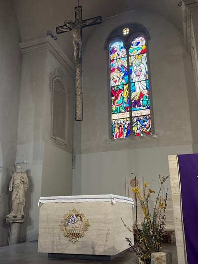 The image depicts the interior of the Duomo di San Feliciano (Cathedral of San Feliciano) in Foligno, Italy. The view is directed towards the main altar and a section of the chancel.
A large, ornate crucifix, featuring the figure of Jesus Christ, is suspended from the ceiling in the upper-left quadrant. The cross itself is dark with gilded or decorative elements along the vertical beam. To the upper right, a tall, arched stained-glass window displays multiple vibrant religious scenes in panels dominated by blue, red, yellow, and green hues.
In the lower-center, a rectangular altar, constructed of light-colored stone (possibly travertine), is covered by a white linen cloth with thin blue and purple stripes. The front of the altar is adorned with a prominent gold-colored circular relief sculpture, depicting a lamb and other symbolic elements.
To the far left, a light-colored stone statue, identifiable as Saint Peter, stands on a decorative pedestal, holding keys in his left hand. A small white security camera is mounted on the upper-left wall below the crucifix. To the right of the altar, a smaller stone lectern or ambo with a microphone stand is visible. Adjacent to it, on the floor, is a tall arrangement of bare branches with small yellow blossoms and green foliage. A vertical purple banner or cloth is partially visible on the far right.
The surrounding walls are light-colored plaster, showing some subtle textural variations and featuring architectural moldings and a decorative recessed niche on the upper-left wall. The floor is a dark, smooth surface.