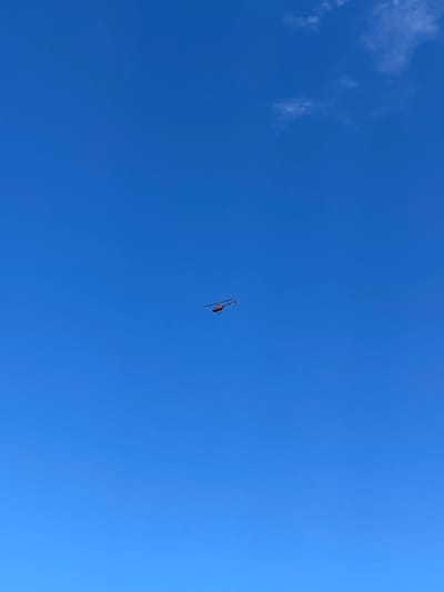 A lone red helicopter is flying in a clear blue sky over Santa Monica, California. The helicopter is in the center of the frame, with a small wisp of cloud in the upper right corner. The helicopter appears to be a small, single-engine model, with its rotor blades extended. The sky is a vibrant blue, with no other visible aircraft or objects. The image is a high-angle shot, looking up at the helicopter from below.