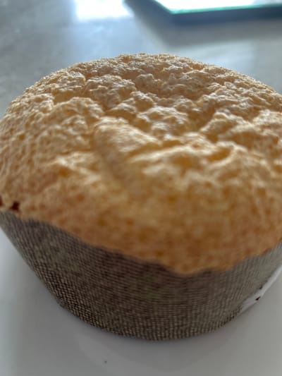 This is a close-up, eye-level, vertical shot of a single, golden-brown muffin with a very porous and uneven top surface. The muffin is encased in a dark, textured paper liner, possibly a baking cup. The muffin is positioned on a white surface, which appears to be a plate. The background is blurred and out of focus, showing hints of a gray surface with a bright light source in the upper portion, and a dark rectangular object in the upper right corner. The scene is indoors. There are no people, animals, or any other objects visible in the image. The image does not contain any elements that would allow for the determination of its specific location within Richmond Hill, Canada.