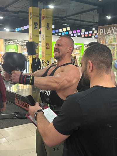 In an indoor environment, likely a shopping mall or recreational center given the colorful suspended lights and large banner with the word "PLAY" visible, a muscular man is performing a weightlifting exercise. He is holding a large, black dumbbell with both hands, his face contorted in exertion. He wears a black tank top and wrist wraps. To his right, another man, with dark hair and a beard, is observing him, holding a clipboard and a phone, possibly acting as a judge or coach. The background shows other people, decorative pillars with cat imagery, and retail displays. The flooring is tiled. The scene appears to be part of a public event or competition.