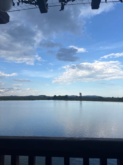 The image shows a waterscape viewed from what appears to be a porch or balcony. To the left is a cloudy sky with varying shades of blue. To the right is a body of water that reflects the sky. In the background, there's a distant treeline and possibly some land. In the foreground on the left, there's a dark structure with metal elements. On the right, is a dark wood frame with rectangular cutouts. Given the information, it is possible this photo was taken in the vicinity of Begunovtsi, Bulgaria, near a lake or river. There is no evidence of people in the frame.