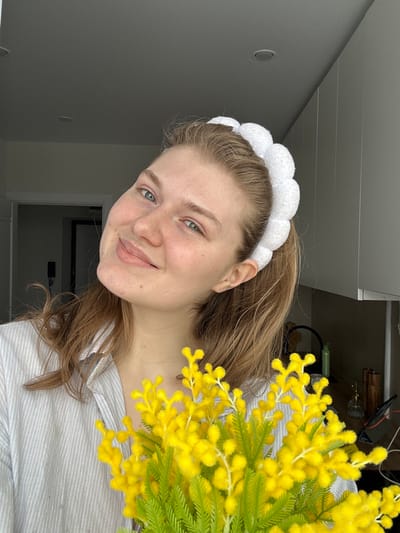 A young Caucasian female subject is depicted from the chest up, positioned indoors in a residential setting in Nikol'skii, Russia. Her light-colored hair is pulled back, secured by a white, puffy, cloud-shaped headband. She looks directly towards the viewer with a subtle smile, exhibiting light-colored eyes. She is wearing a light-colored, possibly striped, collared shirt. In the immediate foreground, she holds a bouquet of artificial bright yellow flowers resembling mimosa, characterized by numerous small yellow florets and green fern-like leaves. The background on the left features a partial doorway leading into a darkened room. On the right, a segment of a modern kitchen or living area is visible, including white overhead cabinetry and a dark countertop. Objects present on the countertop consist of a green bottle, a metallic gold-colored decorative item, a clear glass bottle, and several black electronic cables. The ceiling is a light gray, fitted with two visible recessed lighting fixtures.