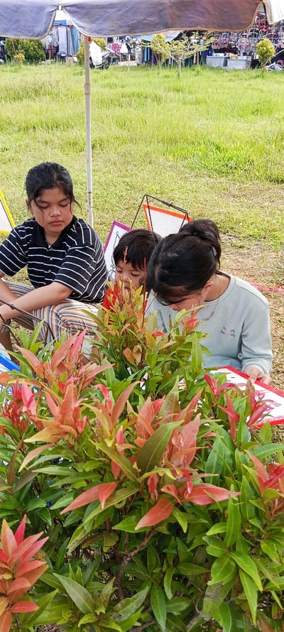 This image, captured in Bandasari, Indonesia, depicts three individuals engaged in an outdoor activity under a grey fabric canopy. The foreground is dominated by a bushy plant featuring green and reddish-orange leaves. To the left, a female with dark hair, wearing a black and white striped polo shirt and striped trousers, is seated on the ground, looking downward. Centrally, a child with dark hair and a red garment is partially visible behind the plant, also directing their gaze downward. To the right, a female with dark hair styled in a ponytail, wearing a light blue long-sleeved shirt with a cat emblem, is bent forward, focused intently on a white board with a red border, held in her left hand, suggesting an activity involving drawing or coloring. Behind the individuals, two black wireframe stands support white boards with yellow and orange trim. The background reveals a grassy field leading to a bustling outdoor market area, characterized by various stalls displaying hanging merchandise, distant indistinct figures, and a visible motorcycle. The scene is illuminated by natural daylight.