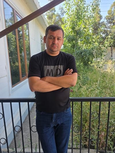 A medium shot of a man standing on a balcony with his arms crossed. He is wearing a black t-shirt with white text and blue jeans. The background is a residential building with large windows and a lush green yard. The image appears to be taken in Karshi City, Uzbekistan, given the architecture and vegetation.