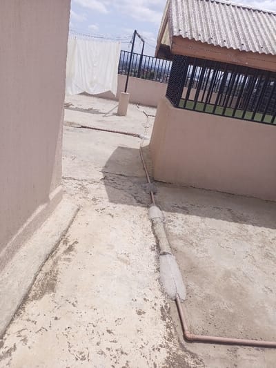 This image captures a utilitarian rooftop scene in Kiamaina ward, Kenya, viewed under a partly cloudy sky. The rooftop surface is a light-colored, rough concrete, exhibiting stains and minor cracking. Two light peach-colored walls delineate a pathway, with one running along the left edge and another forming part of a structure on the right.

A brown/orange pipe, likely conduit or plumbing, extends across the concrete floor, featuring two sections where it is encased in rough, light gray concrete. This pipe then angles towards the right wall. In the mid-ground, a clothesline is strung between a light-colored concrete post and a dark metal pole, supporting multiple white fabric items, presumably laundry, hanging to dry. Behind the clothesline, a dark metal railing provides a vantage point over a distant landscape featuring urban structures.

To the right, an adjacent building structure is visible, topped with a corrugated metal roof. Its upper perimeter features dark metal security bars or a railing, behind which a green surface can be seen. The base of this structure matches the light peach color of the other walls. No individuals are present in the scene.