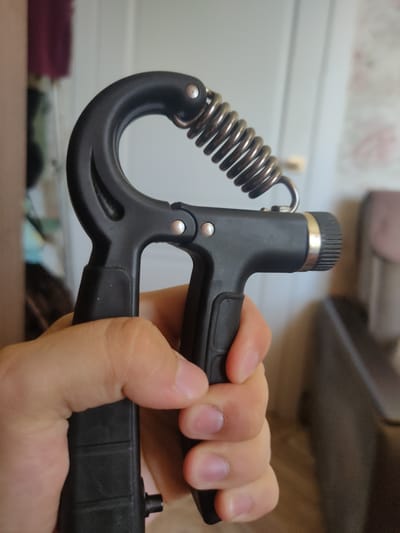 A person's hand is holding a black hand grip exerciser. The exerciser has a curved, black handle with a metal spring connecting the two parts. The hand is gripping the lower part of the exerciser. The background is blurred, but appears to be an indoor setting with a door and wallpaper. The location of Salavat, Russia is not depicted in this image.