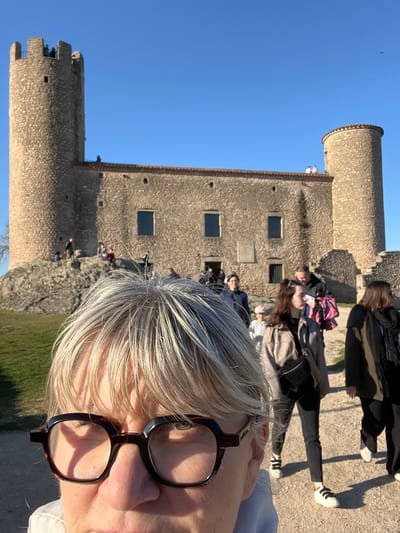 The image depicts an outdoor scene at the Château de Chambles in Chambles, France, under a clear blue sky. In the immediate foreground, the top portion of a person's head is visible, featuring light blonde to grey hair with bangs and dark, square-framed eyeglasses with a tortoise-shell pattern.
The midground shows a dirt path occupied by several pedestrians. A woman with dark hair, dressed in a light beige jacket, dark pants, and white sneakers, walks from left to right, carrying a black sling bag. Behind her, a man with dark hair carries a pink and purple patterned bag. Multiple other individuals are visible walking or standing on the path and surrounding rocky terrain, including a child wearing a pink hat.
The background is dominated by the stone structure of the Château de Chambles. It comprises a central building with a reddish-brown roofline, constructed from rough-hewn light-colored stone, featuring multiple rectangular windows with dark panes. Two prominent round towers flank the central building. The left tower is tall and cylindrical with crenellated battlements, upon which several individuals are standing. The right tower is shorter, also cylindrical, and topped with a rounded, reddish-brown cap. People are also observed on the roofline between the main building and the right tower. The scene is illuminated by strong sunlight.