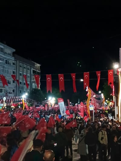 The image shows a large crowd of people gathered in the street in front of a building at night. The crowd is holding red flags with white crescent moons and stars on them. There are several street lights illuminating the scene, and the buildings in the background are lit up. The crowd is largely obscured by the flags, but some individuals are visible, including a few wearing dark clothing. The image appears to have been taken in a public square or on a street in a town in Türkiye, most likely Silivri, based on the provided coordinates. The crowd's density and the flags suggest a celebration or political rally.