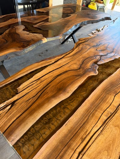 The image depicts a close-up, elevated perspective of a handcrafted wooden table with a unique design featuring a resin river running through its center. The wood grain is prominent, displaying various shades of brown and intricate patterns. The resin portion appears to be a translucent, amber color with a textured surface, mimicking the look of a natural riverbed. The table is supported by black metal legs. The background is blurred, suggesting an outdoor setting with possible construction debris and a reflective surface, and a partial glimpse of what could be a yellow power tool or equipment. There is no indication of the location being Petarch, Bulgaria, or any people present in the scene.