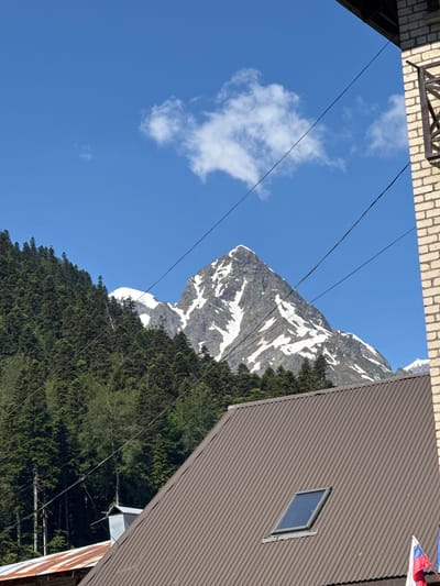 In Dombay, Russia, a mountain scene is captured under a clear blue sky with scattered white clouds. A prominent, snow-capped mountain peak dominates the background, its rugged slopes partially covered in snow. In the foreground, a dense forest of evergreen trees covers a mountainside. A dark brown, ribbed metal roof with a skylight is visible in the lower portion of the frame, partially obscuring the lower slopes of the mountain and forest. To the right, a section of a brick building with a railing is also in view. Several dark wires, possibly power lines or telecommunication cables, crisscross the sky and extend across the image. The presence of the Russian flag suggests a location within Russia.