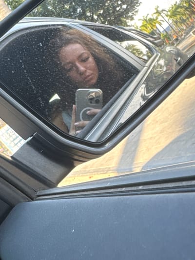 The image shows a woman reflected in a car side mirror. She is holding a smartphone, presumably taking a selfie. The background visible in the mirror suggests an outdoor urban environment with trees and buildings. A street or road can also be seen in the car's mirror. The location is approximated in the city of Lao Cai Ward, Vietnam, but without further evidence to provide confirmation or further details on the environment, this is an approximation.