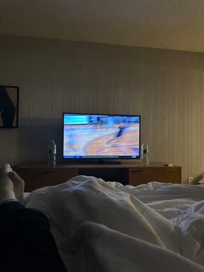 The image captures an indoor scene, likely a bedroom or hotel room, viewed from a low angle, possibly from a bed. The dominant feature is a flat-screen television mounted on a textured, horizontally striped wall in a neutral gray-beige color. To the left, a plain lighter beige wall forms a corner.

The television is displaying what appears to be a live sports broadcast, specifically a basketball game, evidenced by the visible court lines and colors. The image on the screen is slightly blurred, suggesting movement. Visible text on the TV screen includes the "FS1" logo at the top right of the broadcast area. Below this, a vertical information bar displays text: "CBS: Providence at DePaul airing on FS1 and the FOX Sports app," followed by "Program Alert," and then "FS1" and "LIVE" further down.

Two clear plastic water bottles are distinctively placed on the wall around the TV. One is balanced on top of the television, and another is positioned on the wall directly below the TV.

To the right of the television, there is a piece of wooden furniture, possibly a dresser or cabinet, with a dark top surface. Further to the right, a significant portion of white, rumpled bedding is visible, indicating a bed. Partially visible from beneath the bedding are the lower legs and feet of a person, wearing white socks and dark pants or clothing.

At the bottom left corner of the image, a framed item or artwork is partially visible, featuring dark and light blue geometric shapes.

The room is dimly lit, suggesting it is evening or night, with the television screen acting as a primary light source. The overall impression is one of a person relaxing in bed, watching television.
