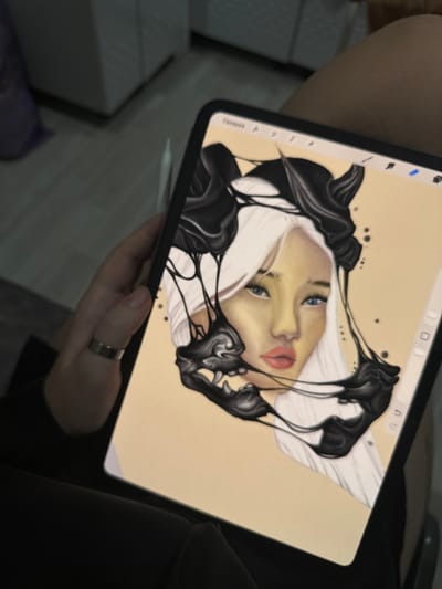 A person is holding a tablet computer which displays a digital illustration of a woman's face with white hair and blue eyes. Black, viscous liquid or slime drips around her face. The tablet is in landscape orientation and rests on the person's lap. The person's left hand, wearing a silver ring, is visible on the left side of the tablet. The background is out of focus and shows what appears to be interior flooring and furniture. There is no discernible connection to Pervouralsk, Russia, in the image.