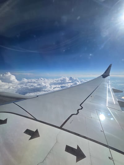 The image shows a view from an airplane window, looking out at the sky and clouds. The airplane's wing is visible in the foreground, with the Ryanair logo clearly displayed on the wingtip. There are also two black arrows painted on the wing, indicating a direction. The sky is a vibrant blue, with scattered white clouds below and to the left. The sun is shining brightly in the upper right corner, causing some lens flare. The overall scene depicts a typical airplane view during flight. There is no information within the image to suggest a specific location in Ghindărești, Romania.