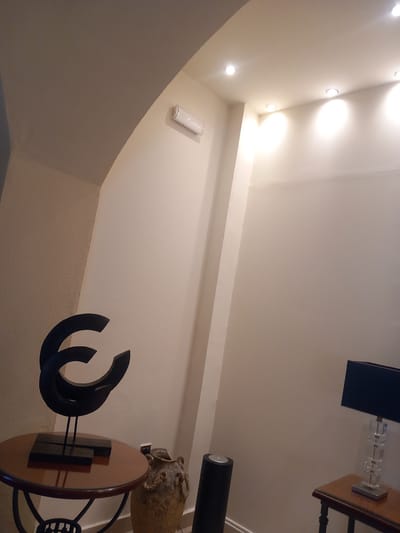 The image depicts an indoor scene within a building, likely a residence or commercial establishment, located in the city of Kos, Greece. The environment appears to be a hallway or an entryway, characterized by curved archways in the ceiling and neutral-toned walls with a textured finish.
Prominently featured is a dark, abstract sculpture with curved elements, positioned on a circular wooden table with a dark rim and a metal base. To the right of the table, a textured, urn-like decorative vase with rope detailing sits on the floor. Further to the right, a cylindrical black object, possibly a speaker or a stand, is visible. In the background, a rectangular table with a wooden top supports a table lamp with a black shade and a clear, faceted base. The lighting in the room is provided by recessed ceiling lights and the table lamp, casting a warm glow on the surfaces. The overall impression is one of modern interior design with a touch of artistic decoration.