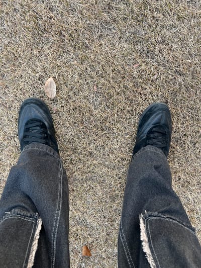 A top-down shot of a person standing on dry, sparse grass. They wear black sneakers and dark gray jeans with frayed hems. One sneaker is near a small, dry leaf, while another leaf is near the other sneaker. The jeans are cuffed, and the overall impression is casual and slightly worn.