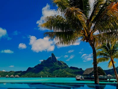 The scene depicted does not represent a location within Lillestrøm, Norway. Instead, it features a tropical island landscape under a clear blue sky with scattered white cumulus clouds. In the foreground, an infinity pool with deep blue water reflects the sky. To the right, two palm trees, one large and central, the other smaller, frame the scene. The midground consists of a calm, turquoise lagoon. On the left, a series of overwater bungalows with thatched roofs are visible on stilts. To the right, a wooden pier extends into the water, connecting to a larger thatched-roof structure. A white leisure boat is docked alongside this structure. Several small white sailboats are scattered across the water in the mid-distance. The background is dominated by a large, lush green volcanic mountain with a distinct jagged peak, characteristic of Mount Otemanu, rising from the island. No individuals, explicit actions, or interactions are observable.
