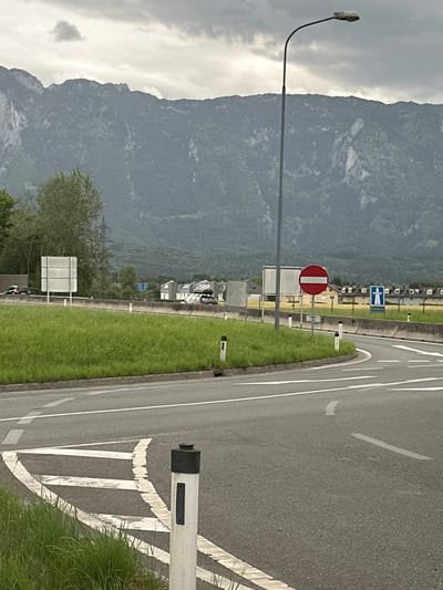 The image depicts a gray, overcast day in Walserfeld, Austria, with a mountain range in the background. The foreground shows a curving asphalt road with white lane markings and a grassy verge. A white bollard with a black top stands on the right side of the road. In the middle ground, there is a traffic island with a large white sign. Further on, a red circular sign with a white horizontal bar, indicating "No Entry," is visible. Beyond that, a blue sign with a white symbol of a road and an arrow is present. Several buildings with dark roofs are situated in the background, along with some trees and what appears to be a highway. Cars are visible on the road in the middle distance. A tall street lamp stands to the left of the "No Entry" sign.