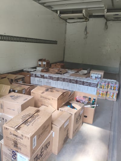 The image shows the interior of a refrigerated truck filled with boxes of alcoholic beverages. The boxes are stacked on top of each other, some are open, revealing bottles inside. The products visible include vodka, cognac, and possibly beer cans. Brands and labels such as "Домашняя", "Старый Какет", "Scotch Terrier", and "LAVETTI" are identifiable. The environment appears to be a loading bay or storage area. The presence of refrigeration units at the top of the truck indicates the goods are being transported under controlled temperature conditions. There are no people visible in the image. The scene is a still, matter-of-fact depiction of inventory in a transport vehicle.