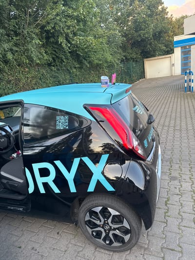 The image shows the rear driver's side of a black car with light blue lettering and accents parked on a paved surface. The car has a QR code on the rear passenger window and the lettering "ORYX" is visible on the side. There are two cans on the roof of the car. In the background, there are trees, a garage door, and a building with blue and white stripes. The scene appears to be in a commercial or industrial area. There are no people visible in the image. The location is confirmed to be Ahlen, Germany.