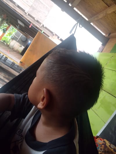 A close-up, side profile shot shows a young child lying in a dark fabric hammock. The child's head is turned away from the camera, revealing the back of their head and neck. Their hair is short and appears wet. The child is wearing a dark t-shirt with a white collar and some white text on the front, partially obscured by the hammock. The background is a blur of light and green. Above the child, a wooden structure with a corrugated roof is visible, with ropes and what appears to be a clothesline with a red clothespin hanging from it. The scene suggests an outdoor or semi-outdoor environment, possibly a backyard or porch area.