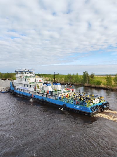 The image shows a blue and white river tugboat, identified as "OT-2452," with a barge attached to its side. The vessel is moving through a dark, possibly muddy river in Ozernoe Ust'e, Russia, as evidenced by the vessel's official website. The barge is loaded with gray material, possibly gravel or sand. The tugboat has multiple decks and a control tower. The stern of the tugboat shows a warning message, "ОСТОРОЖНО-ВИНТ TAKE CARE-SIDEPROPELLER." The river is bordered by green trees and vegetation. The sky above is partly cloudy with patches of blue.