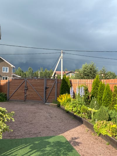 This media file shows a residential property in Kiskelovo, Russia, with a focus on the entrance gate and surrounding garden. The scene depicts a wooden double gate, partially open, with metal framing and latches. Adjacent to the gate is a neatly landscaped garden featuring various green shrubs, flowering plants with light blue and yellow blossoms, and a gravel pathway. A section of a house with a brick facade and white-framed windows is visible to the left of the gate. Power lines with a white utility pole run across the frame, indicating the presence of infrastructure.
The weather appears overcast and possibly stormy, with a dark, dramatic sky dominating the upper left portion of the image. This creates a contrast with the bright green foliage and the well-maintained garden. The lighting suggests it might be late afternoon or early evening, with shadows cast across the gravel and artificial turf to the right of the path. The overall mood is one of a quiet, possibly impending change in weather, over a suburban or rural residential setting. There is no visible text in the image.