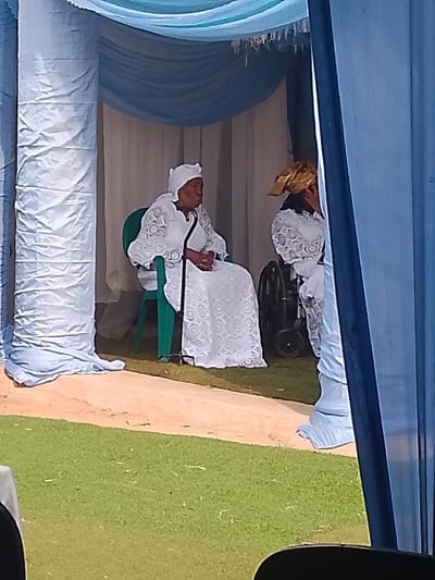 The scene depicts an outdoor or semi-outdoor event in Ufuma, Nigeria, characterized by ceremonial fabric decorations. Two individuals are seated under a canopy draped with light blue and darker blue fabrics.

On the left, an elderly woman is seated upright in a dark green plastic chair. She wears a white headwrap and a long-sleeved white dress with a patterned lace overlay featuring concentric circles. Her hands are clasped in her lap. A black walking stick rests against her left side, extending to the ground. She gazes towards the right, out of frame.

To her right, a second individual is partially visible, seated in a black wheelchair. This person also wears a white garment with a similar patterned lace and a gold-colored headwrap. The back wheel of the wheelchair is clearly visible.

The ground consists of a patch of green grass where the chairs are placed, bordered by a lighter-colored, possibly sandy or dirt path in the foreground. Large structural columns, wrapped in light blue fabric, flank the seating area. A prominent dark blue fabric drape on the far right partially obscures the scene. The overhead canopy structure is further adorned with a light-colored, fringed fabric element.